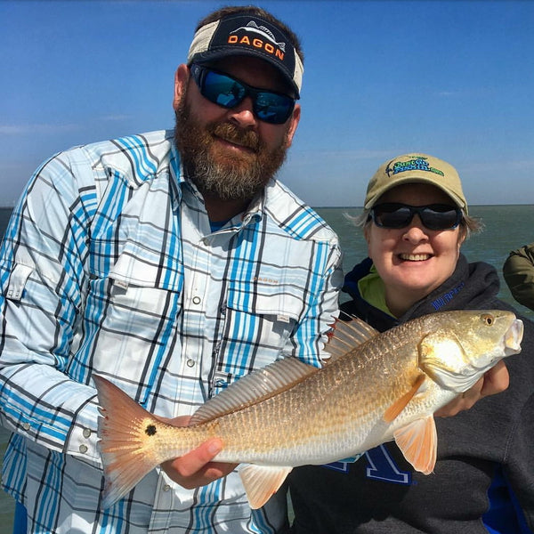 Man wearing Dagon fishing visor and madras blue shirt with a catch in hand, and a woman in background.
