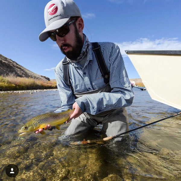 An angler with Navy Gingham Shirt, a wader, and hat catches a fish. 